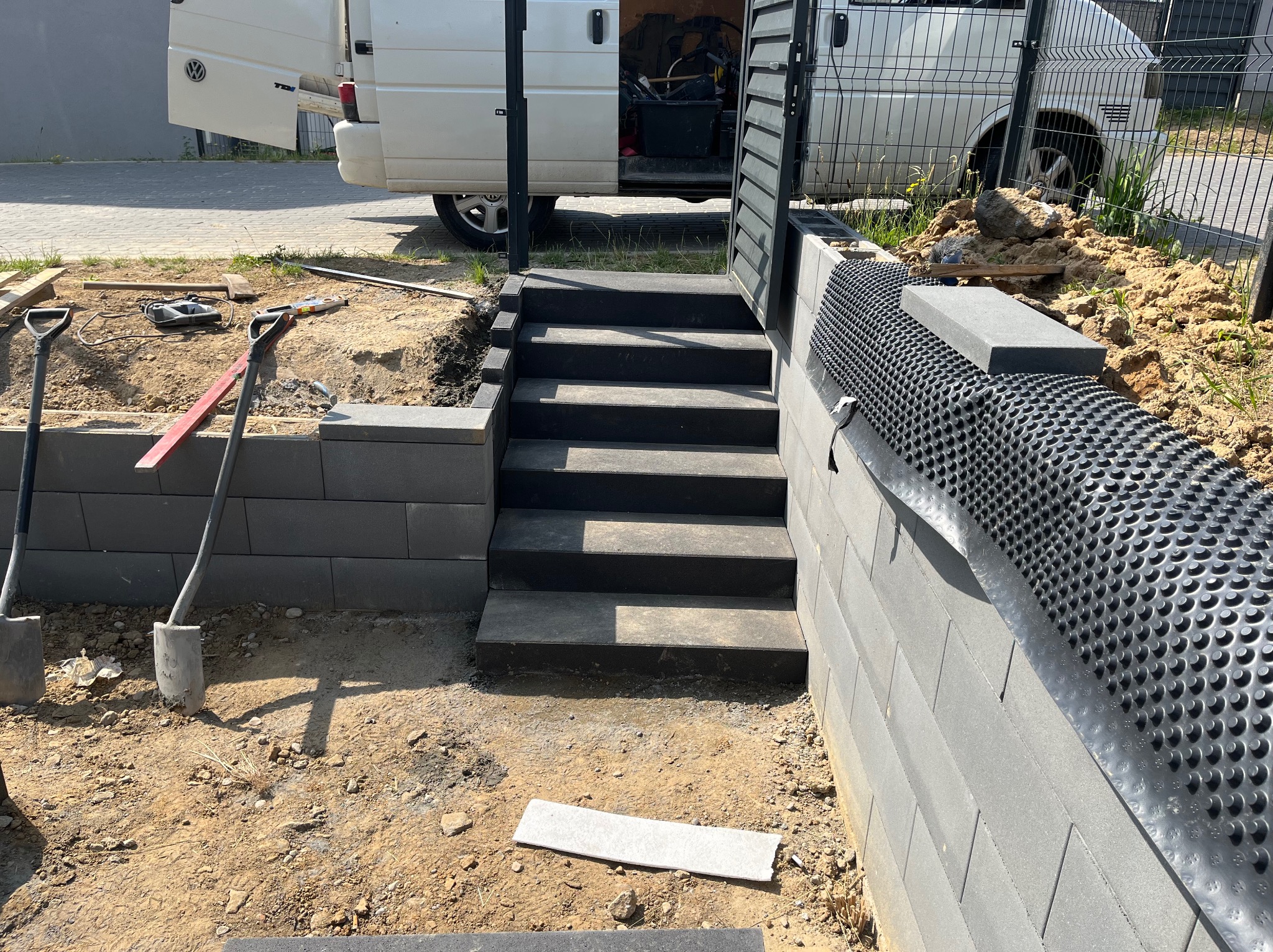 Budowa schodów zewnętrznych z betonowych elementów, widoczne narzędzia i materiały budowlane, w tle częściowo otwarty van.