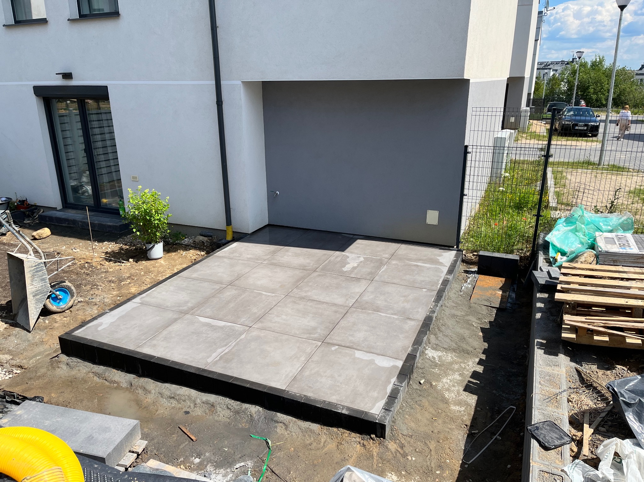 Ułożona, szara, kwadratowa nawierzchnia z kostki brukowej przy nowoczesnym domu z szarą bramą garażową, widoczne narzędzia brukarskie i ziemia do wyrównania terenu.
