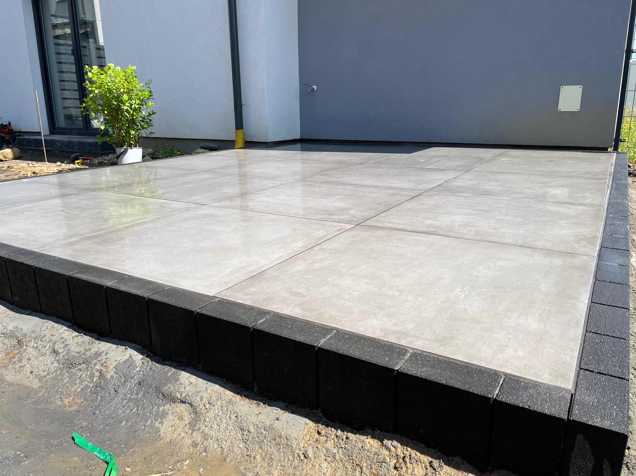 Nowoczesny taras wyłożony jasnoszarą, kwadratową płytką z ciemnoszarą obwódką z kostki; widoczne ślady wilgoci po myciu, jasne ściany budynku w tle.