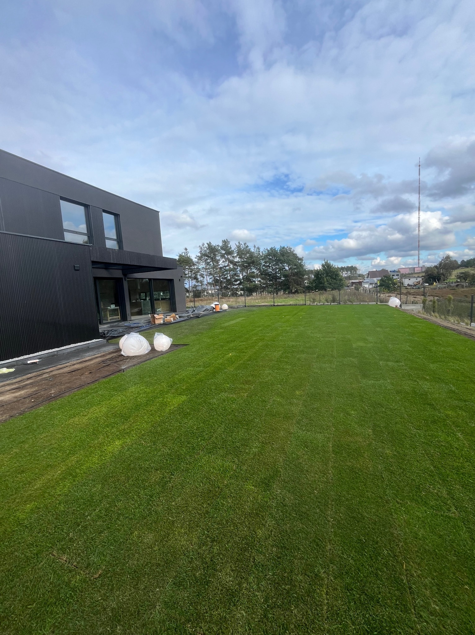 Nowo założony trawnik z rolki przed nowoczesnym domem z czarną elewacją. Widoczne białe worki i przygotowane podłoże. W tle drzewa i maszt telekomunikacyjny.