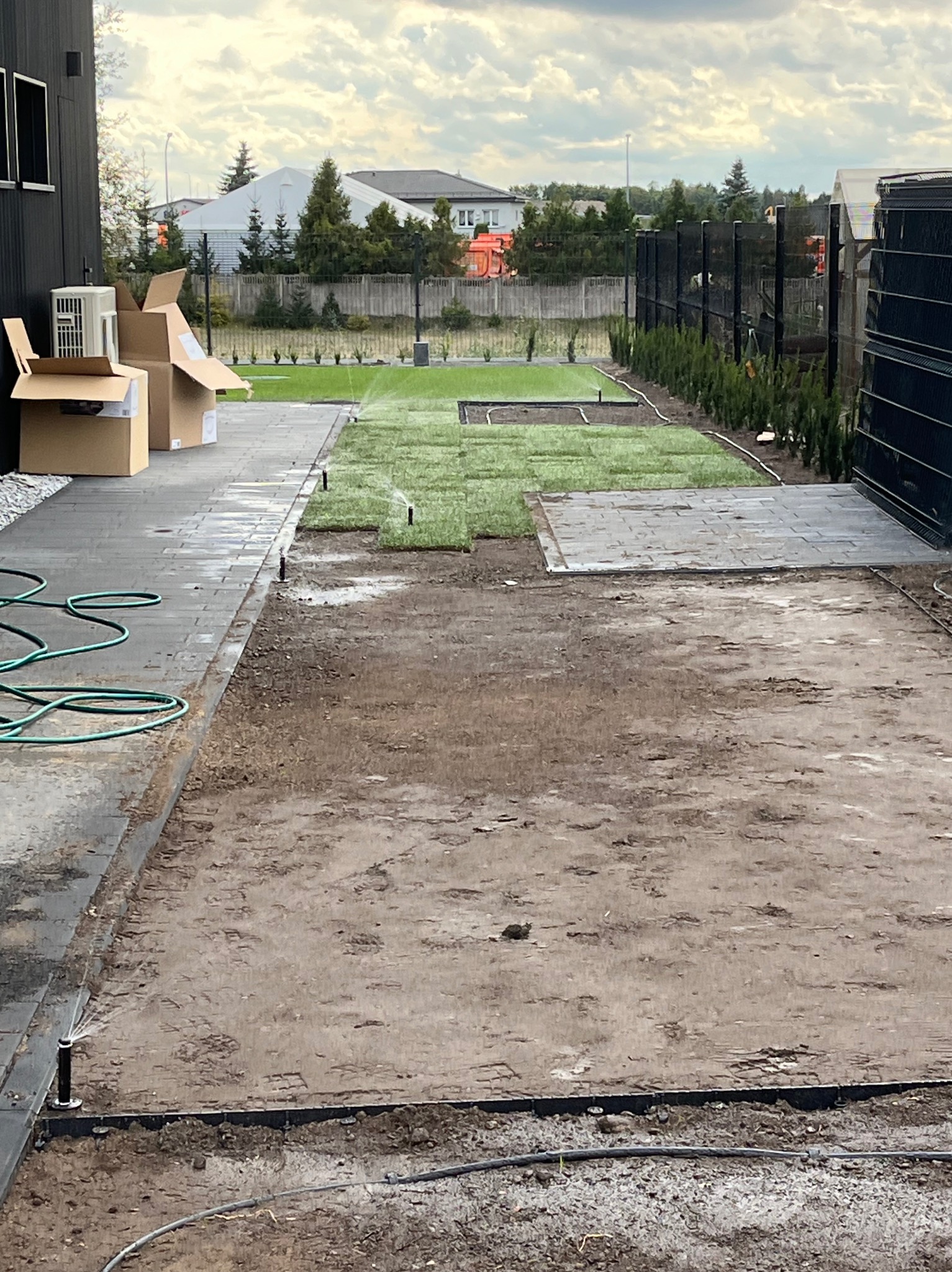 Układanie trawy z rolki i montaż systemu nawadniania w ogrodzie, widoczne rozłożone trawniki, system zraszaczy, przygotowany teren pod trawnik, kostka brukowa i ogrodzenie panelowe w tle.