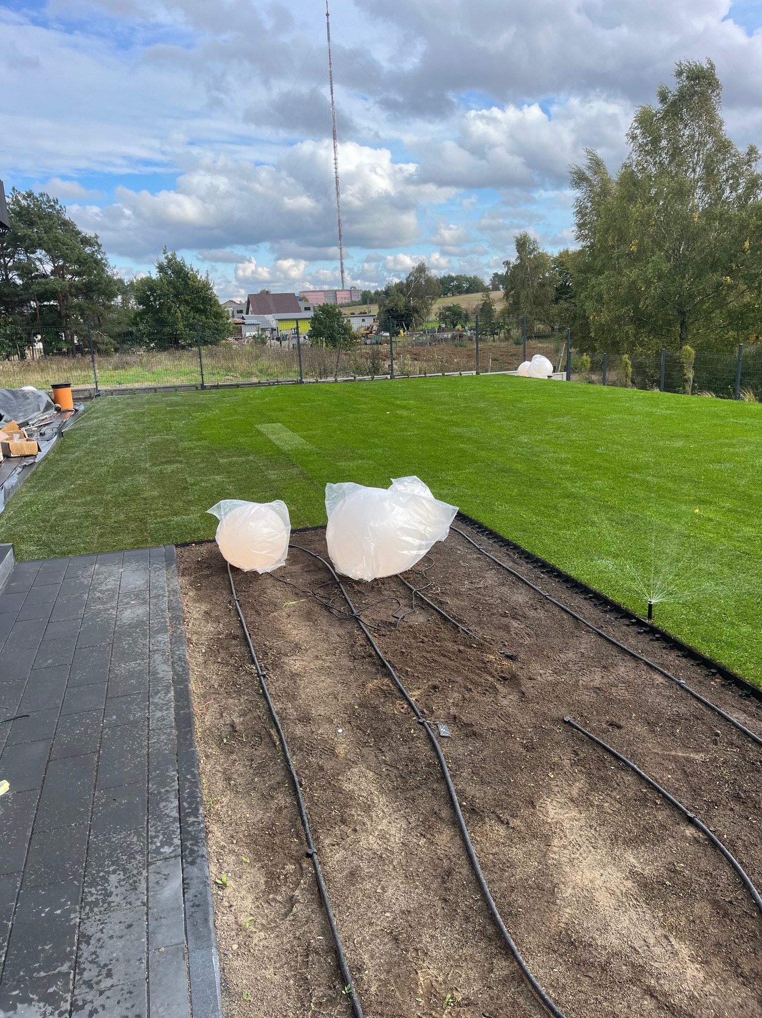 Świeżo ułożona trawa z rolki w Gdańsku, widoczne elementy systemu nawadniania w trakcie instalacji oraz przygotowane do montażu lampy ogrodowe w foliowych osłonach.