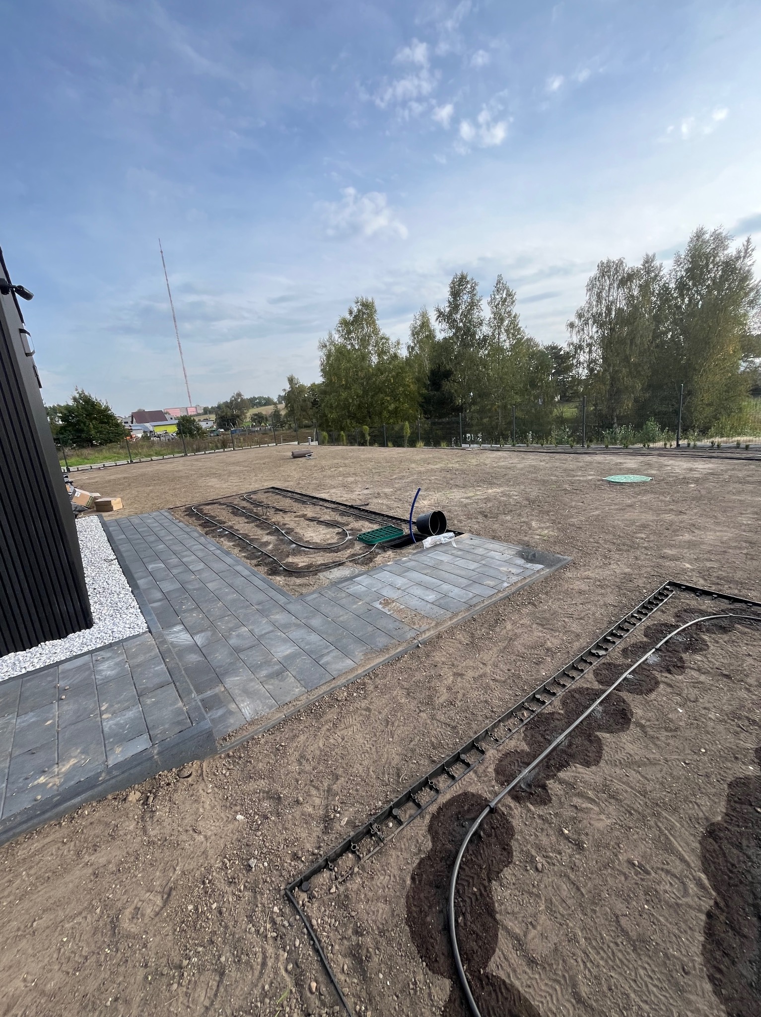 Przygotowanie terenu pod trawnik z rolki: ułożona kostka brukowa, system nawadniania w trakcie instalacji, widoczne rury i zraszacze, ziemia przygotowana pod trawnik, w tle drzewa i zabudowania.