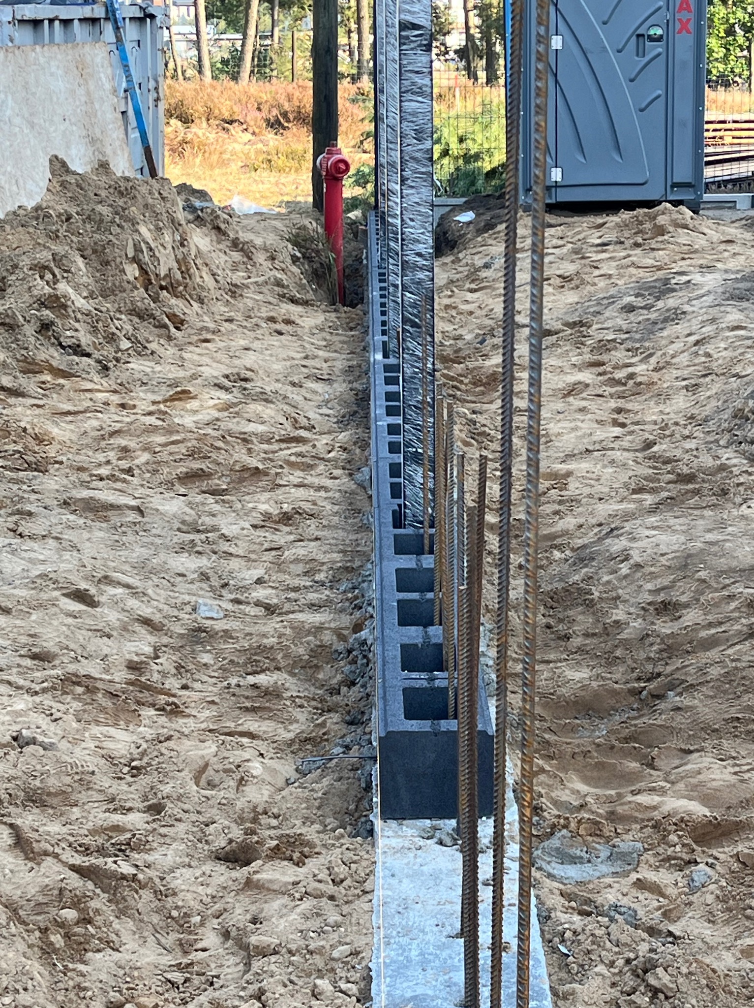Wykop z ułożonymi bloczkami betonowymi tworzącymi fundament pod ogrodzenie, z wystającymi prętami zbrojeniowymi, na tle ziemi i hydrantu.