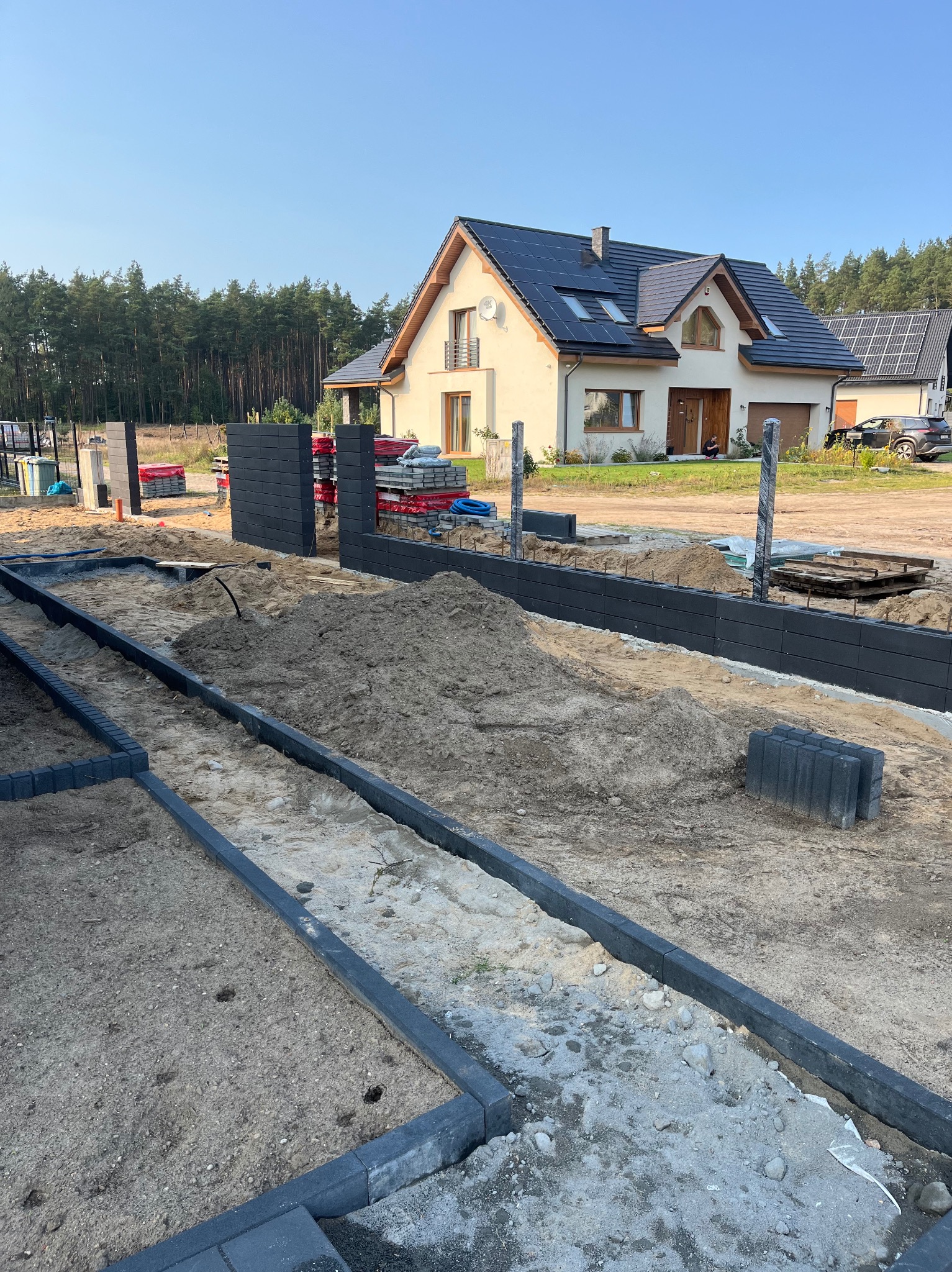 Wykop pod fundament ogrodzenia z betonowych paneli, widoczne przygotowane słupki, w tle dom jednorodzinny z panelami słonecznymi.