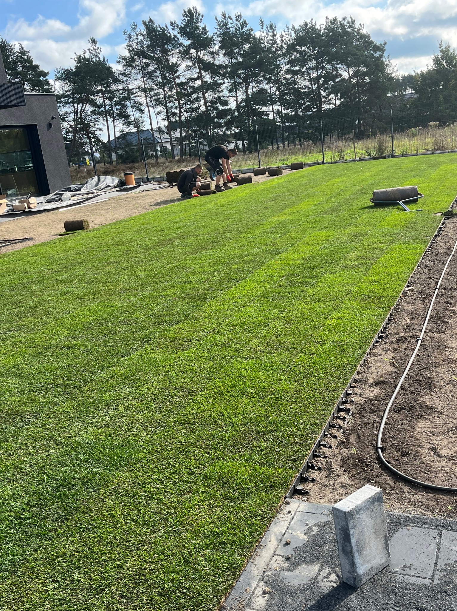 Dwaj mężczyźni układają rolki trawy na przygotowanym podłożu, widoczny trawnik, wał do trawy, granica trawnika i nowoczesny dom w tle.