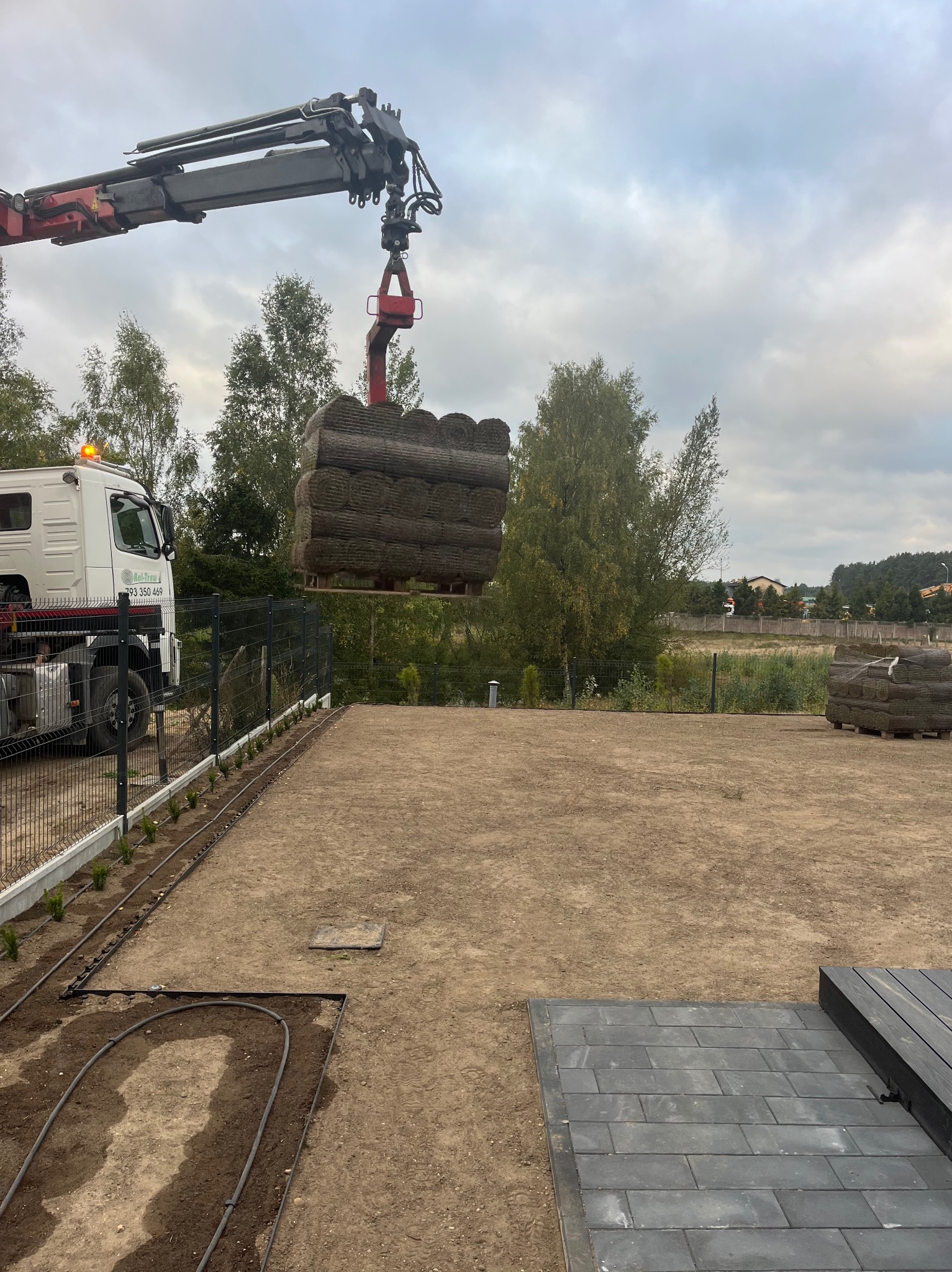 Dostawa trawy z rolki na palecie za pomocą dźwigu samochodowego na przygotowany teren z widoczną instalacją nawadniającą i fragmentem ułożonej kostki brukowej.
