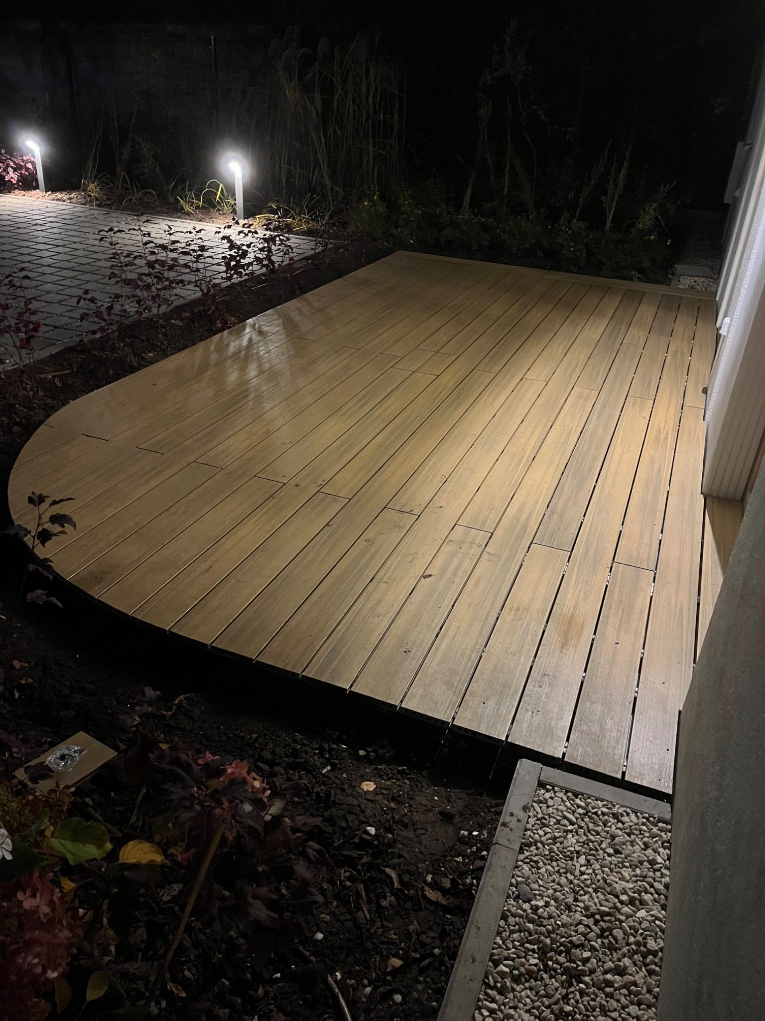 Nowoczesny taras z desek kompozytowych o zaokrąglonym kształcie, oświetlony lampami ogrodowymi, wykonany w ciemnych tonacjach.