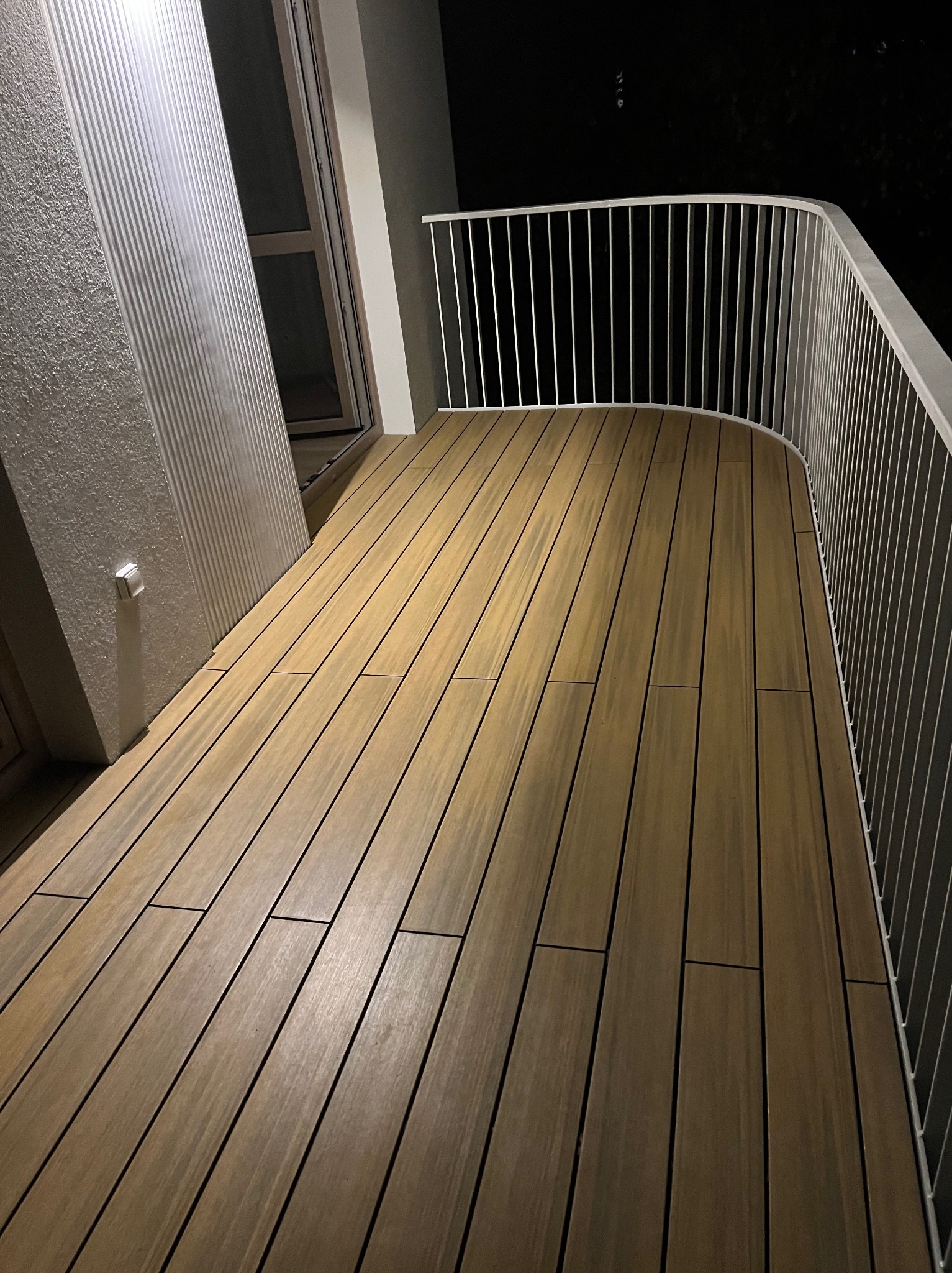 Nowoczesny taras z desek kompozytowych w odcieniach brązu, zabezpieczony białą, metalową balustradą, oświetlony delikatnym światłem.