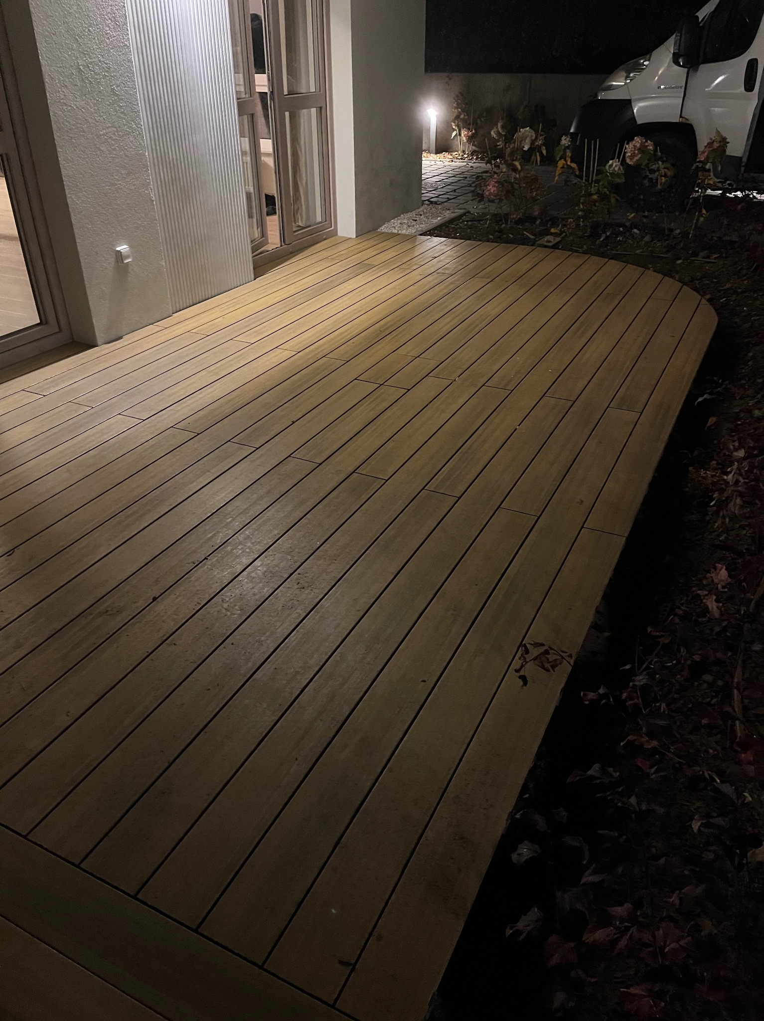 Nowoczesny taras z desek kompozytowych w odcieniach brązu, oświetlony nocą, z widocznym fragmentem ogrodu i zaparkowanym samochodem w tle.