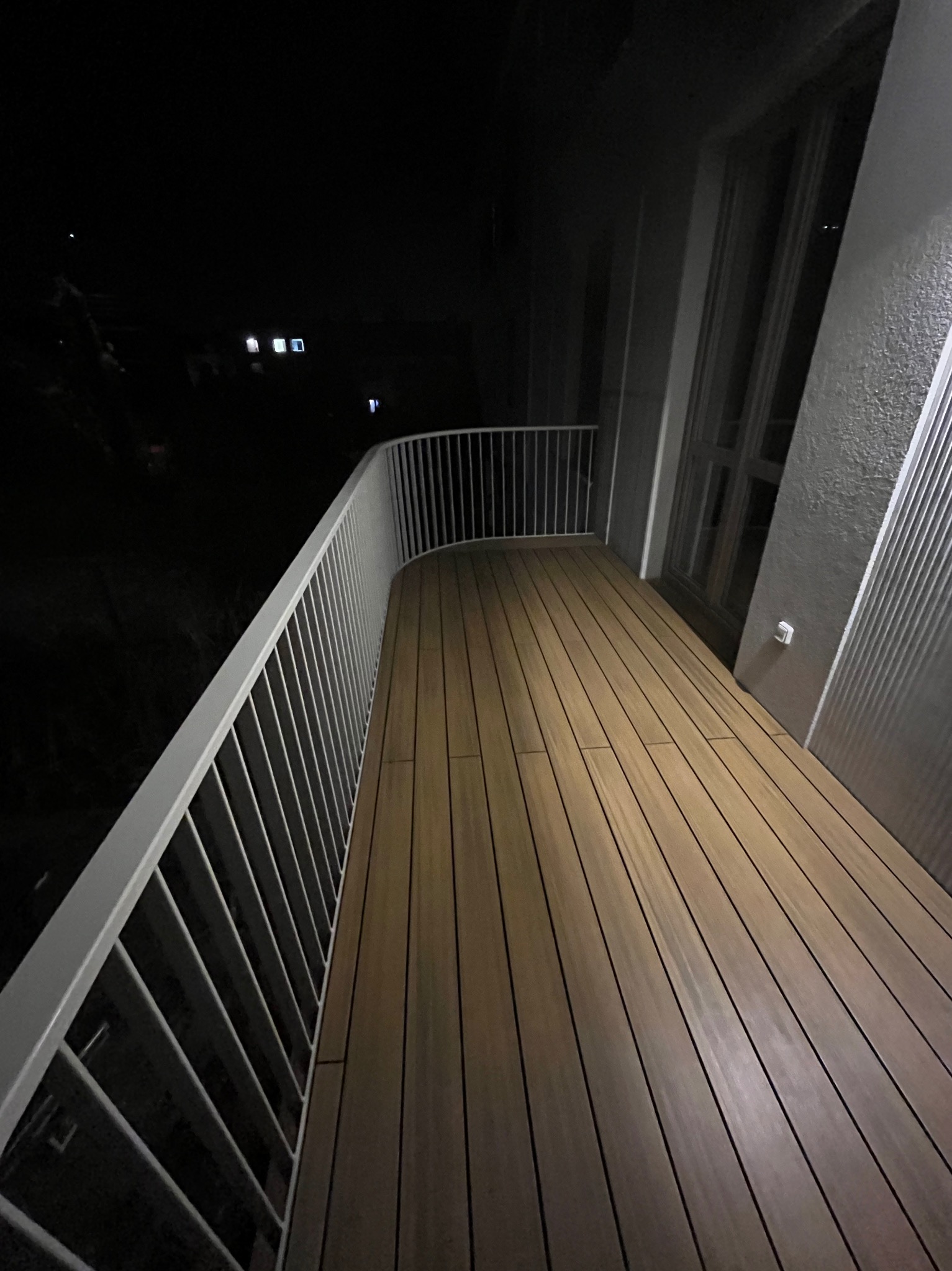 Nocne ujęcie balkonu z drewnianą podłogą i białymi, metalowymi barierkami, widok na ciemny krajobraz z odległymi światłami.