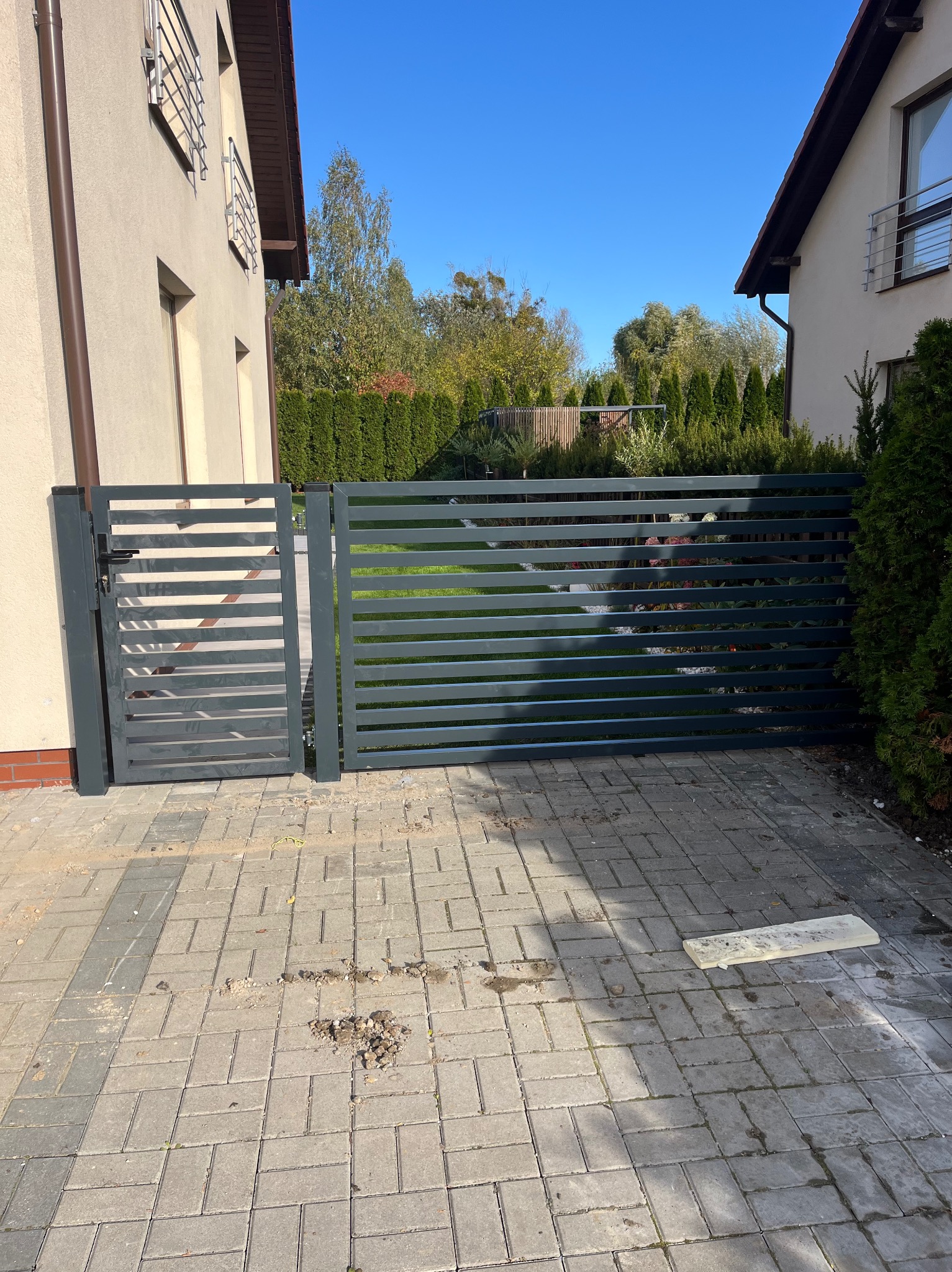 Nowoczesna, grafitowa brama posesyjna z poziomych profili, widok z zewnątrz na kostkę brukową i zieleń ogrodu w tle.