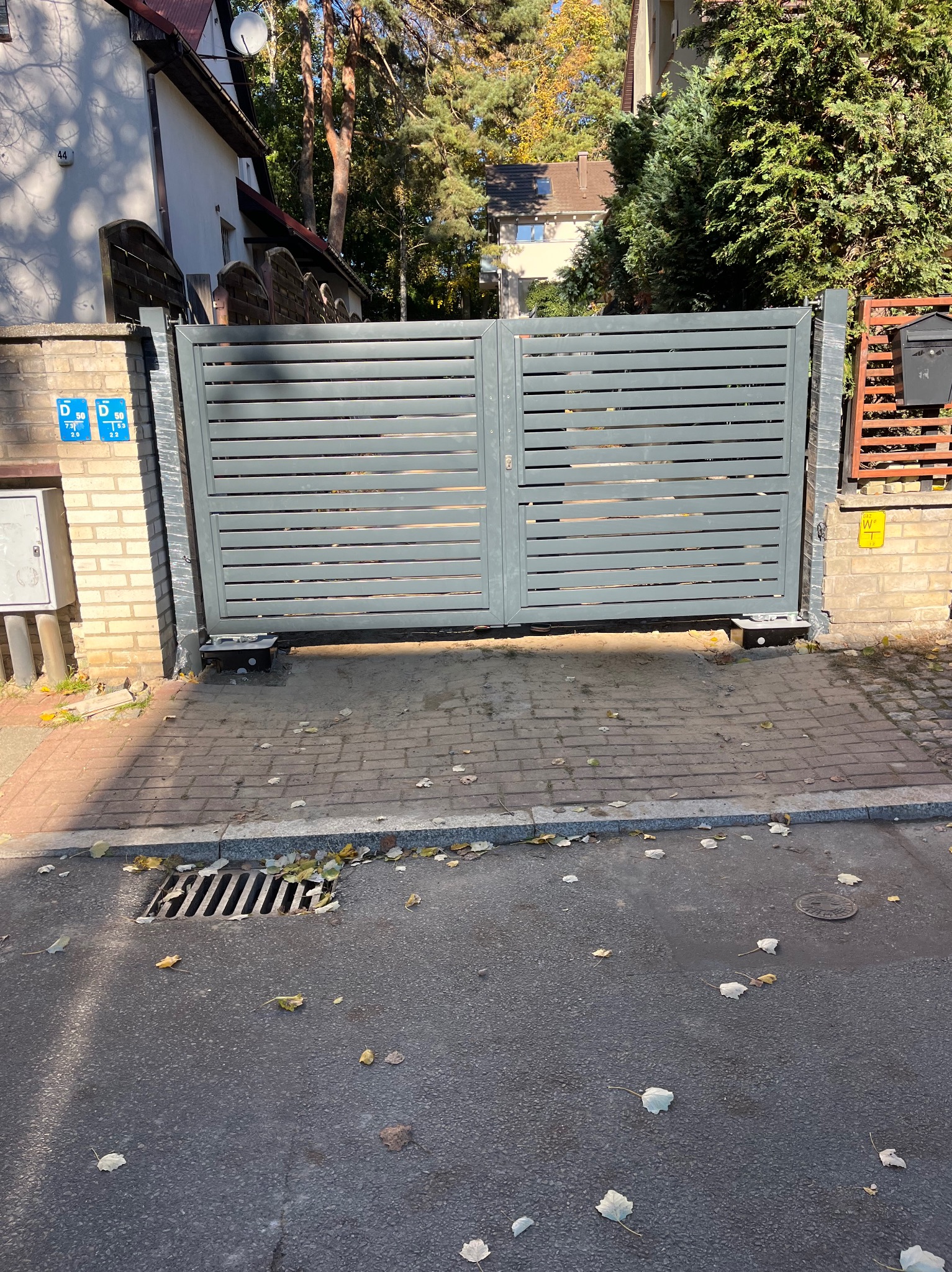 Nowoczesna, szara brama przesuwna z poziomych paneli, zamontowana na ceglanych słupkach, widok z zewnątrz na chodnik i ulicę z opadłymi liśćmi.