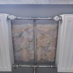 prosuccess.sp.z.o.o - Dwa białe grzejniki podłączone chromowanymi rurami, widoczne zawory termostatyczne, w tle ściana z resztkami starej, kwiatowej tapety.