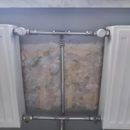 Dwa białe grzejniki podłączone chromowanymi rurami, widoczne zawory termostatyczne, w tle ściana z resztkami starej, kwiatowej tapety.