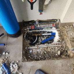 prosuccess.sp.z.o.o - Otwór w podłodze z widocznymi rurami instalacji grzewczej i hydraulicznej w trakcie prac remontowych, fragmenty styropianu i gruzu na podłodze, niebieski filtr wody obok.