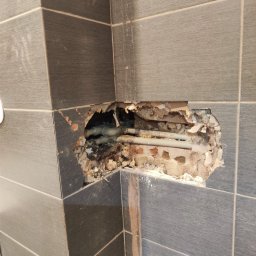 prosuccess.sp.z.o.o - Odkryte rury wodne w ścianie z ciemnych płytek, uszkodzona ściana wokół rur, widoczny gruz i fragmenty cegieł.