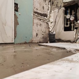 prosuccess.sp.z.o.o - Niedokończona instalacja hydrauliczna: widoczne rury, liczniki, piana montażowa, oraz mokra posadzka w trakcie prac remontowych.