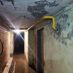 prosuccess.sp.z.o.o - Żółta rura gazowa biegnąca wzdłuż ściany w piwnicy z łuszczącą się farbą, widoczna drabina i oświetlenie w głębi korytarza.