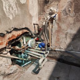 prosuccess.sp.z.o.o - Fragment ściany z widoczną instalacją hydrauliczną w trakcie naprawy, rury z zaworami, wodomierz i narzędzia hydrauliczne na podłodze z płytek.