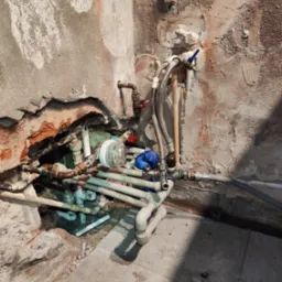 Fragment ściany z widoczną instalacją hydrauliczną w trakcie naprawy, rury z zaworami, wodomierz i narzędzia hydrauliczne na podłodze z płytek.