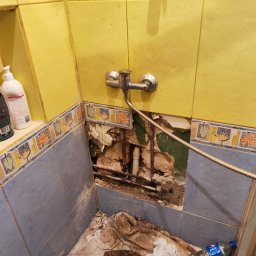 prosuccess.sp.z.o.o - Odkryte rury w ścianie kabiny prysznicowej z usuniętymi płytkami, widoczny kran, resztki gruzu i butelka wody na podłodze.