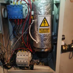 Wnętrze skrzynki z piecem CO owiniętym folią aluminiową z naklejką ostrzegawczą o wysokim napięciu, widoczne przewody, bezpieczniki i elementy instalacji hydraulicznej.