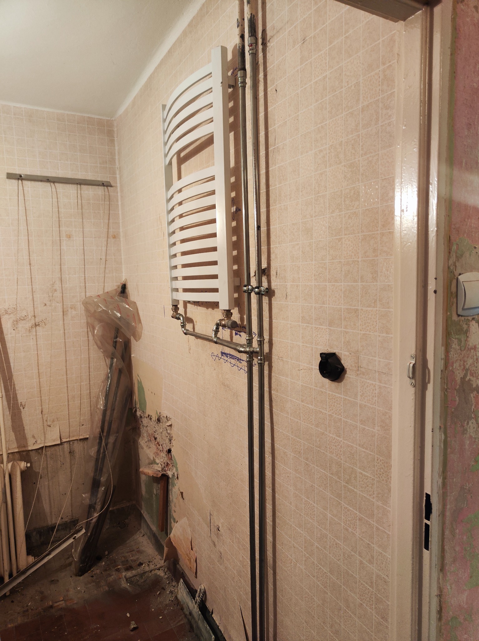 Biały, drabinkowy grzejnik łazienkowy zamontowany na ścianie z beżowych płytek w trakcie remontu; widoczne rury z zaworami, uszkodzone fragmenty ściany i pozostałości po starej instalacji.