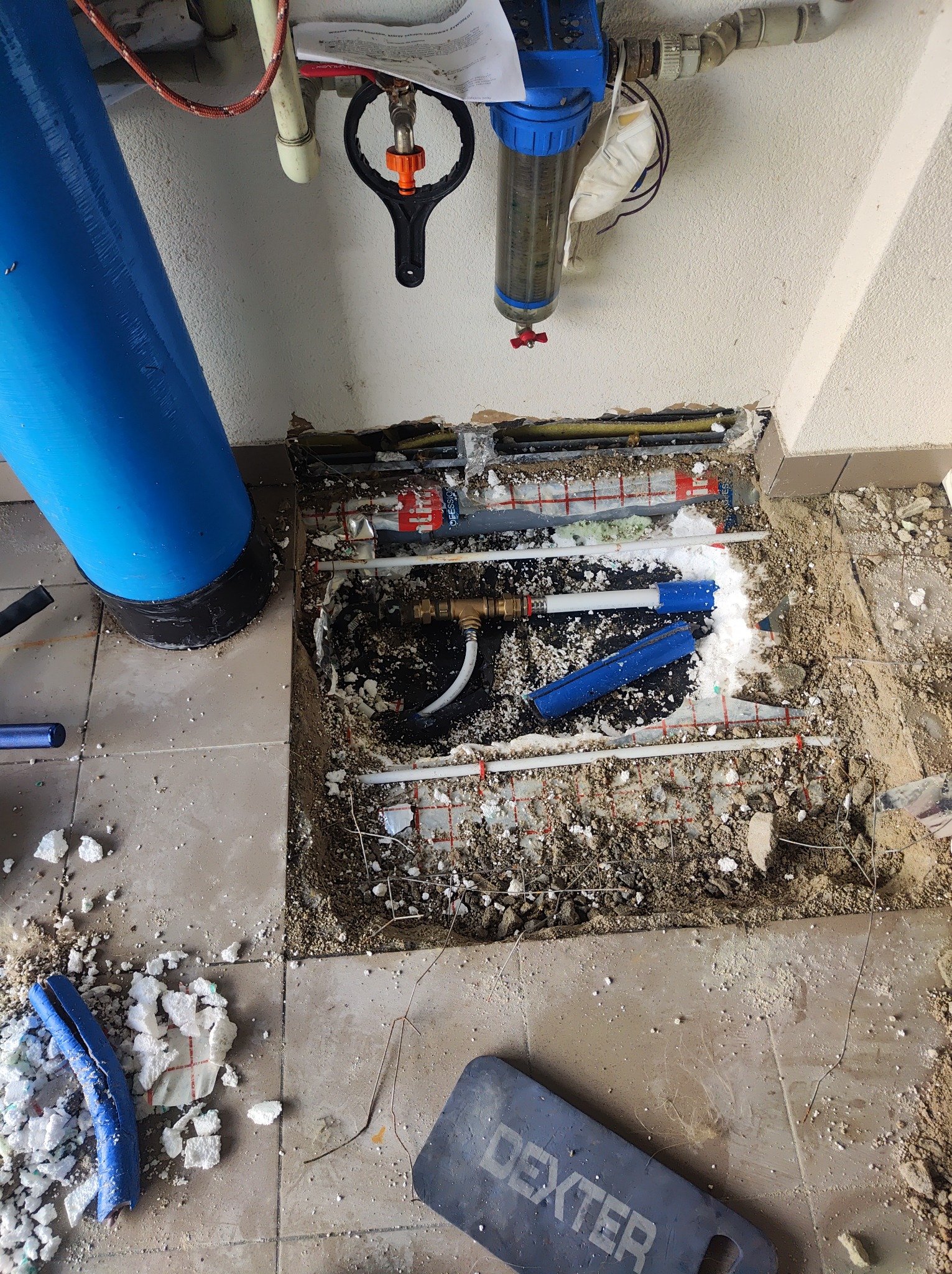 Otwór w podłodze z widocznymi rurami instalacji grzewczej i hydraulicznej w trakcie prac remontowych, fragmenty styropianu i gruzu na podłodze, niebieski filtr wody obok.