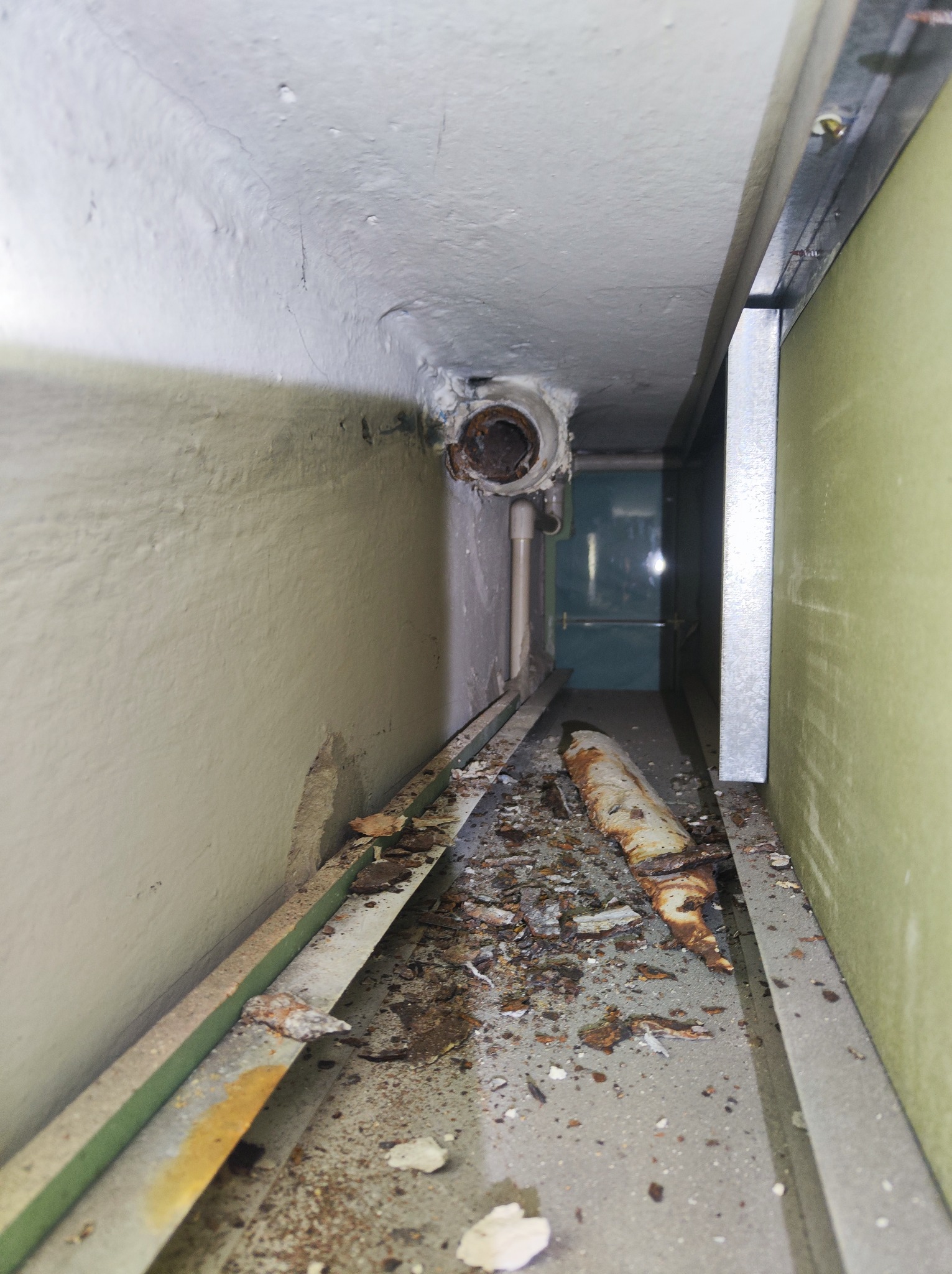 Wąski kanał instalacyjny z widoczną rurą, fragmentami starej izolacji i rdzą na podłodze, wnętrze sprawia wrażenie zaniedbanego i wymaga interwencji hydraulicznej.