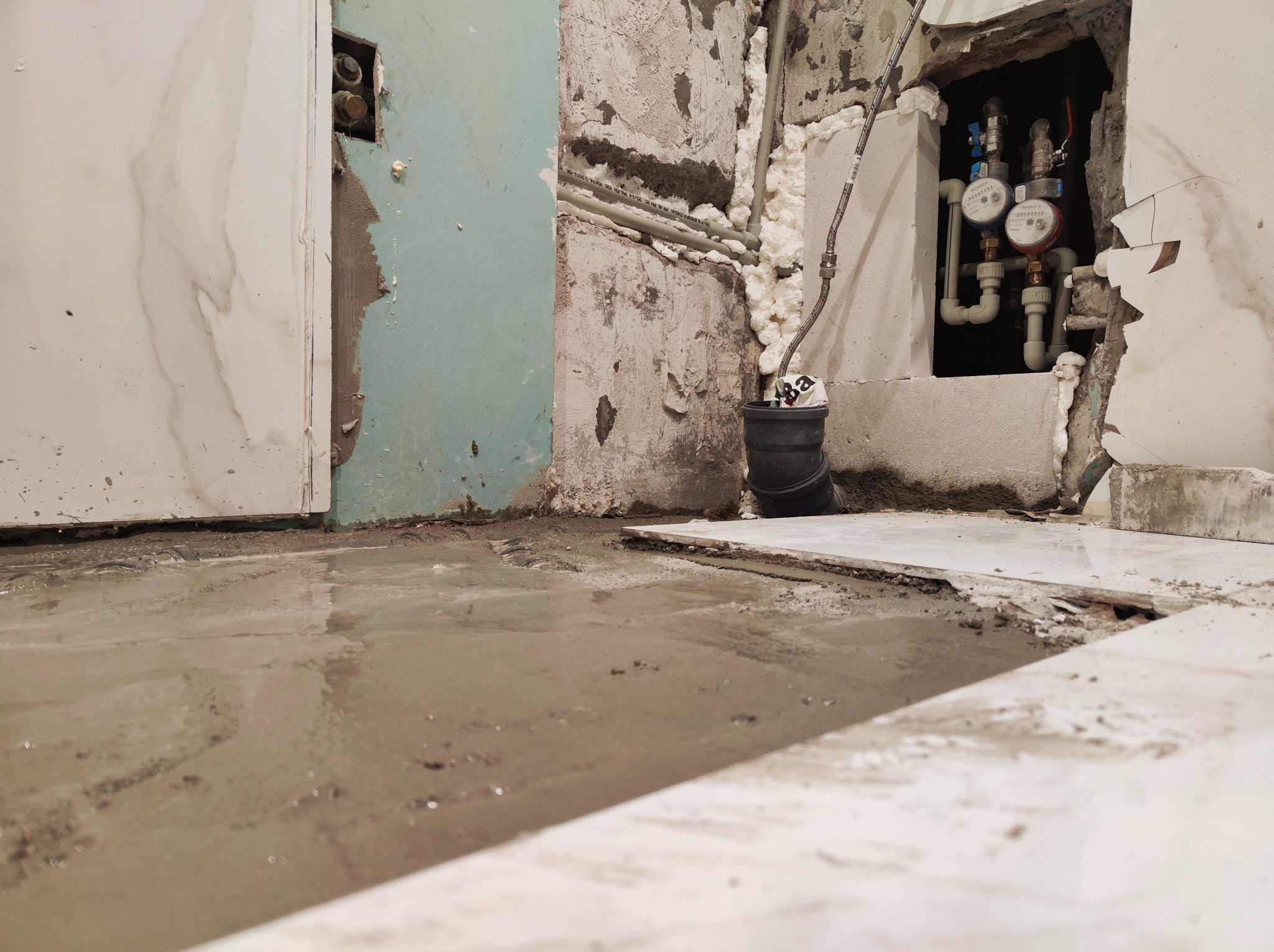 Niedokończona instalacja hydrauliczna: widoczne rury, liczniki, piana montażowa, oraz mokra posadzka w trakcie prac remontowych.