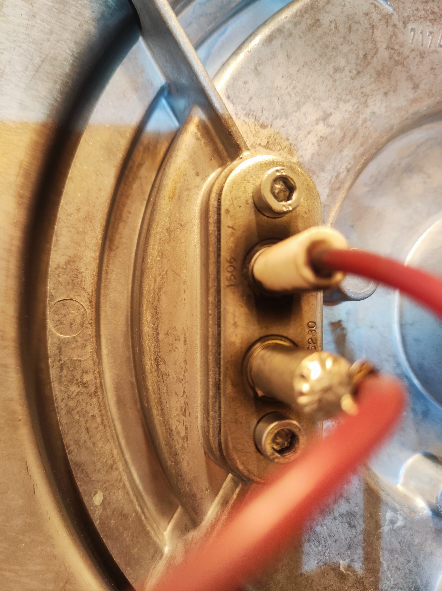 Zbliżenie na metalowy element pieca z dwoma śrubami, podłączonymi przewodami i numerami seryjnymi, widoczne ślady użytkowania.