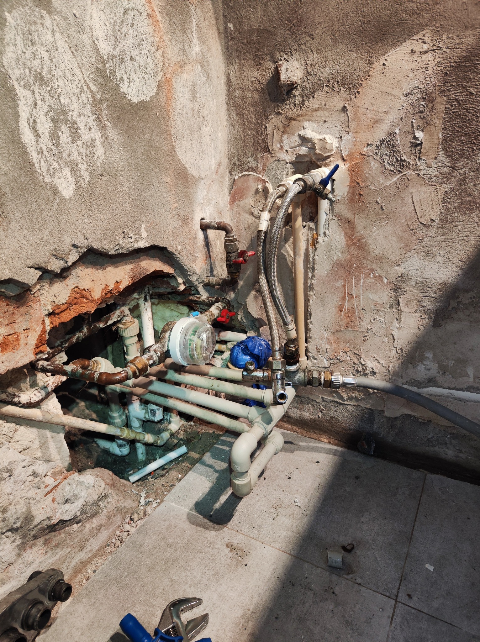 Fragment ściany z widoczną instalacją hydrauliczną w trakcie naprawy, rury z zaworami, wodomierz i narzędzia hydrauliczne na podłodze z płytek.