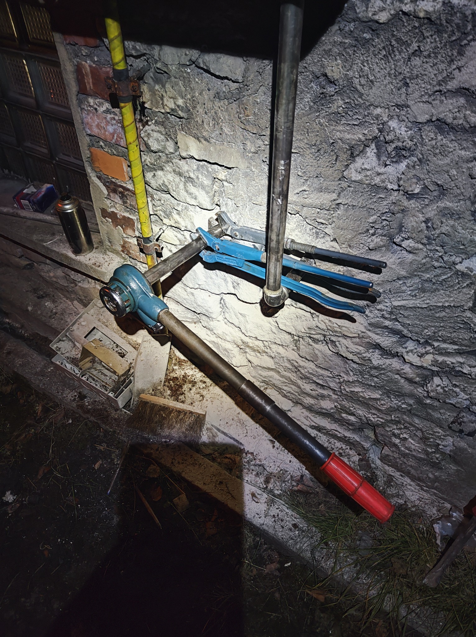 Narzędzia hydrauliczne, w tym gwintownica z długą rękojeścią i niebieskie klucze nastawne, leżące na tle surowej ściany z kamienia i cegły w trakcie prac instalacyjnych.
