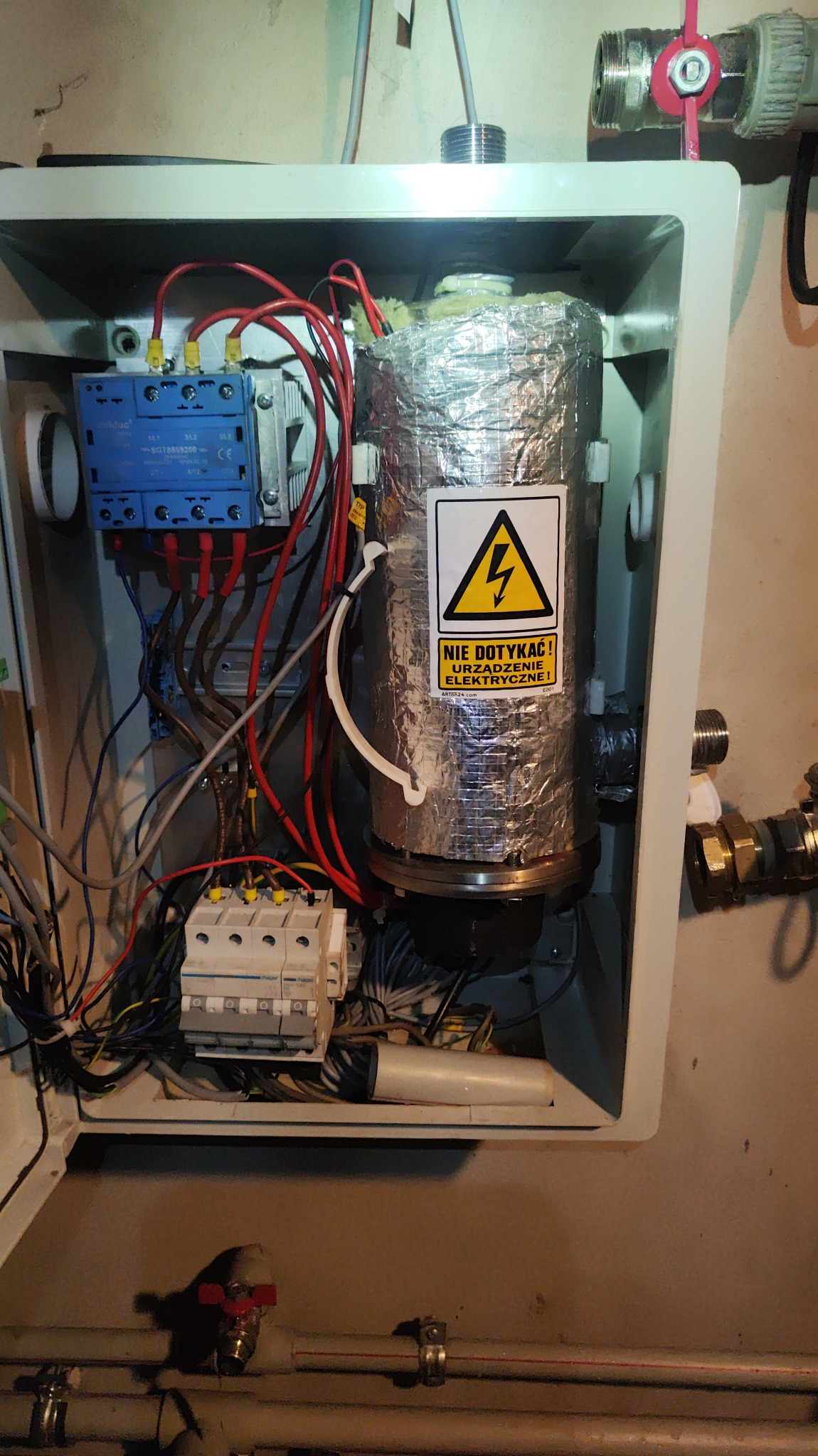 Wnętrze skrzynki z piecem CO owiniętym folią aluminiową z naklejką ostrzegawczą o wysokim napięciu, widoczne przewody, bezpieczniki i elementy instalacji hydraulicznej.