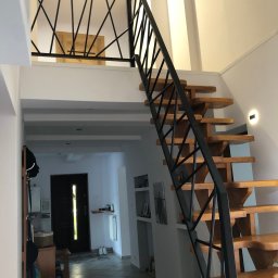 Usługi spawalnicze Mateusz Rodziewicz - Drewniane schody z czarną, geometryczną konstrukcją metalową, prowadzące na antresolę z podobną balustradą, widoczne w jasnym wnętrzu domu.