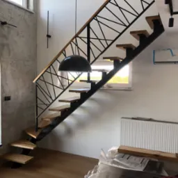 Nowoczesne schody z drewnianymi stopniami i metalową, geometryczną balustradą, oświetlone wiszącą lampą w kształcie kopuły, sfotografowane podczas montażu - widoczne narzędzia i osoba robiąca zdjęcie.