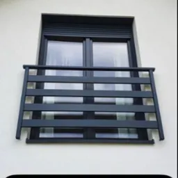 Okienna konstrukcja zabezpieczająca w postaci metalowej balustrady z poziomych profili, zamontowana na zewnątrz budynku z jasną elewacją, widok z dołu.