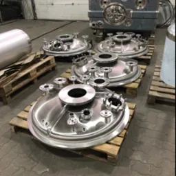 Polerowane, stalowe elementy zbiorników ciśnieniowych na drewnianych paletach w hali produkcyjnej, widoczne różne rozmiary i konfiguracje przyłączy.