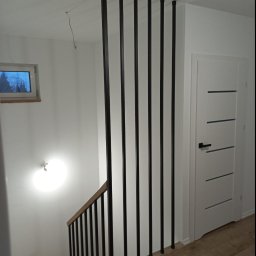 Usługi spawalnicze Mateusz Rodziewicz - Wnętrze klatki schodowej z drewnianymi schodami i balustradą z czarnych, pionowych prętów, widoczna biała ściana z oknem i białe drzwi z poziomymi wgłębieniami, oświetlenie sufitowe.