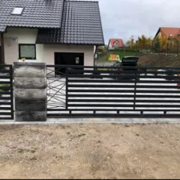 Nowoczesna, przesuwna brama wjazdowa z czarnymi, poziomymi panelami i betonowymi słupkami, widok z zewnątrz posesji, dom jednorodzinny w tle.