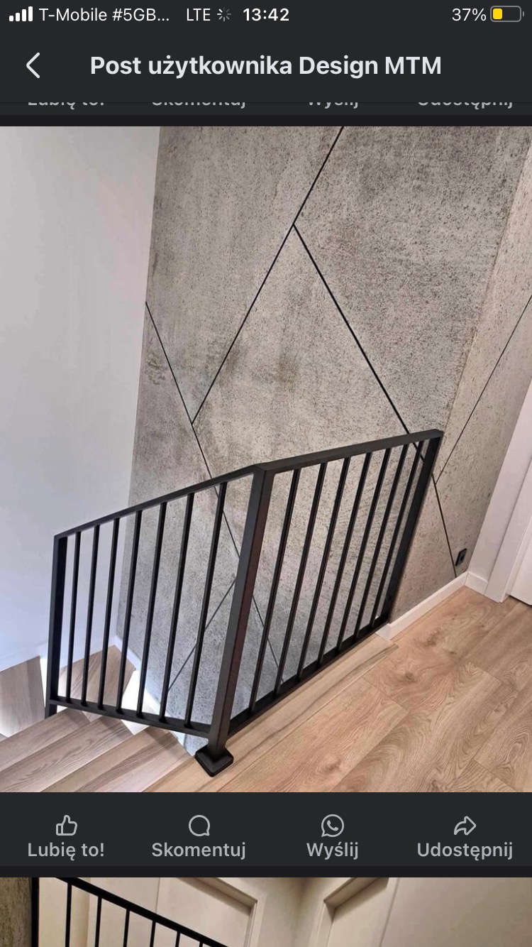 Czarna, metalowa balustrada schodowa z pionowymi szczeblami na drewnianych schodach w Mrągowie, na tle ściany wykończonej płytami o fakturze betonu.