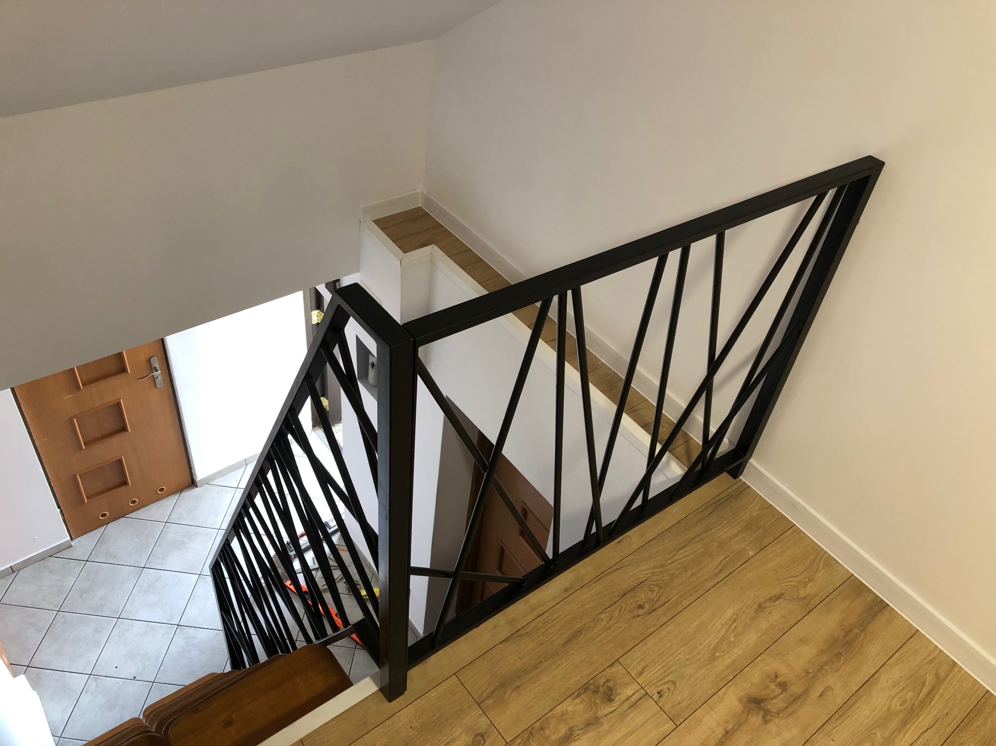 Nowoczesna czarna balustrada schodowa z geometrycznym wzorem, widok z góry na drewniane schody i podłogę, białe ściany.