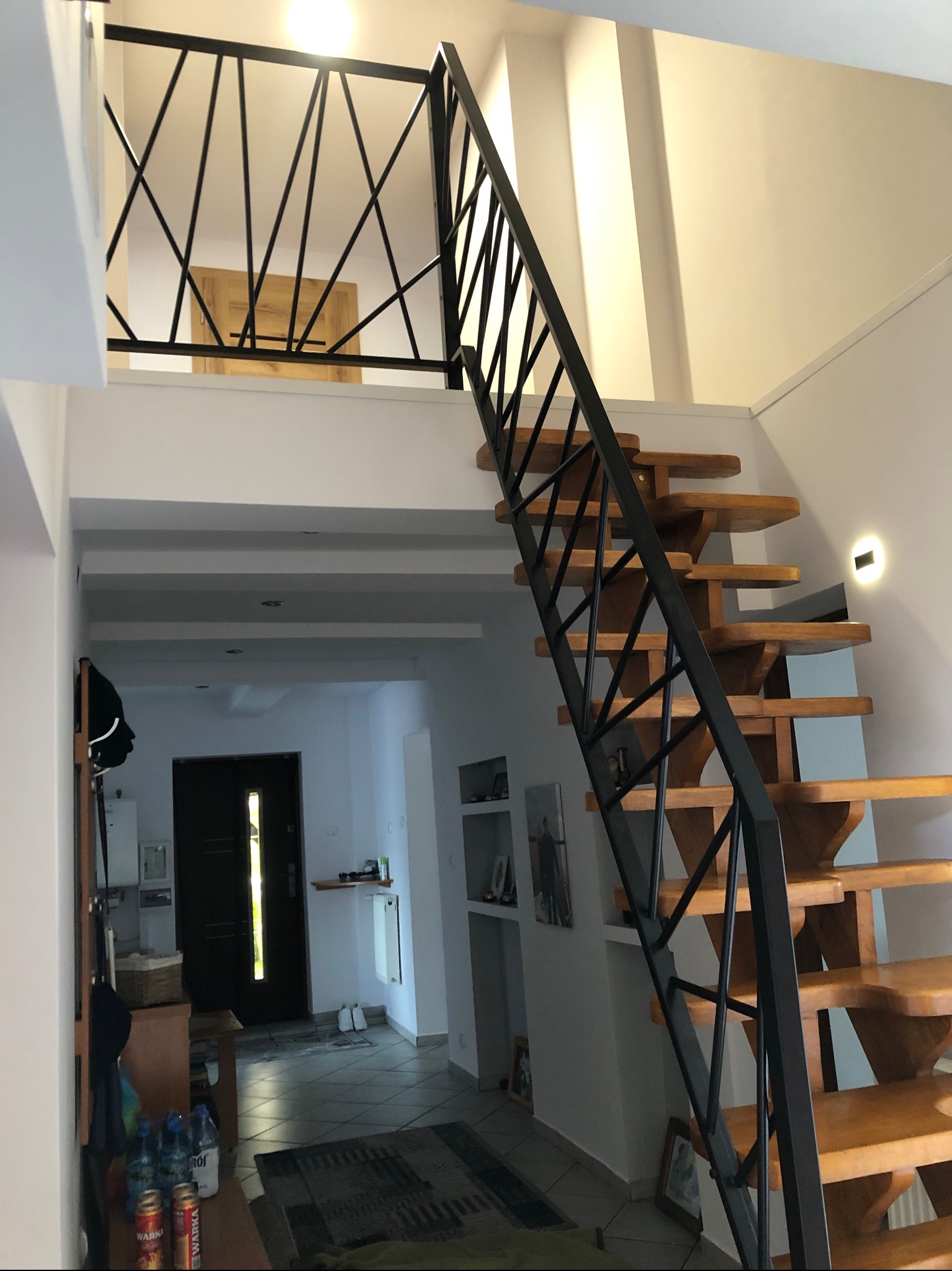 Drewniane schody z czarną, geometryczną konstrukcją metalową, prowadzące na antresolę z podobną balustradą, widoczne w jasnym wnętrzu domu.