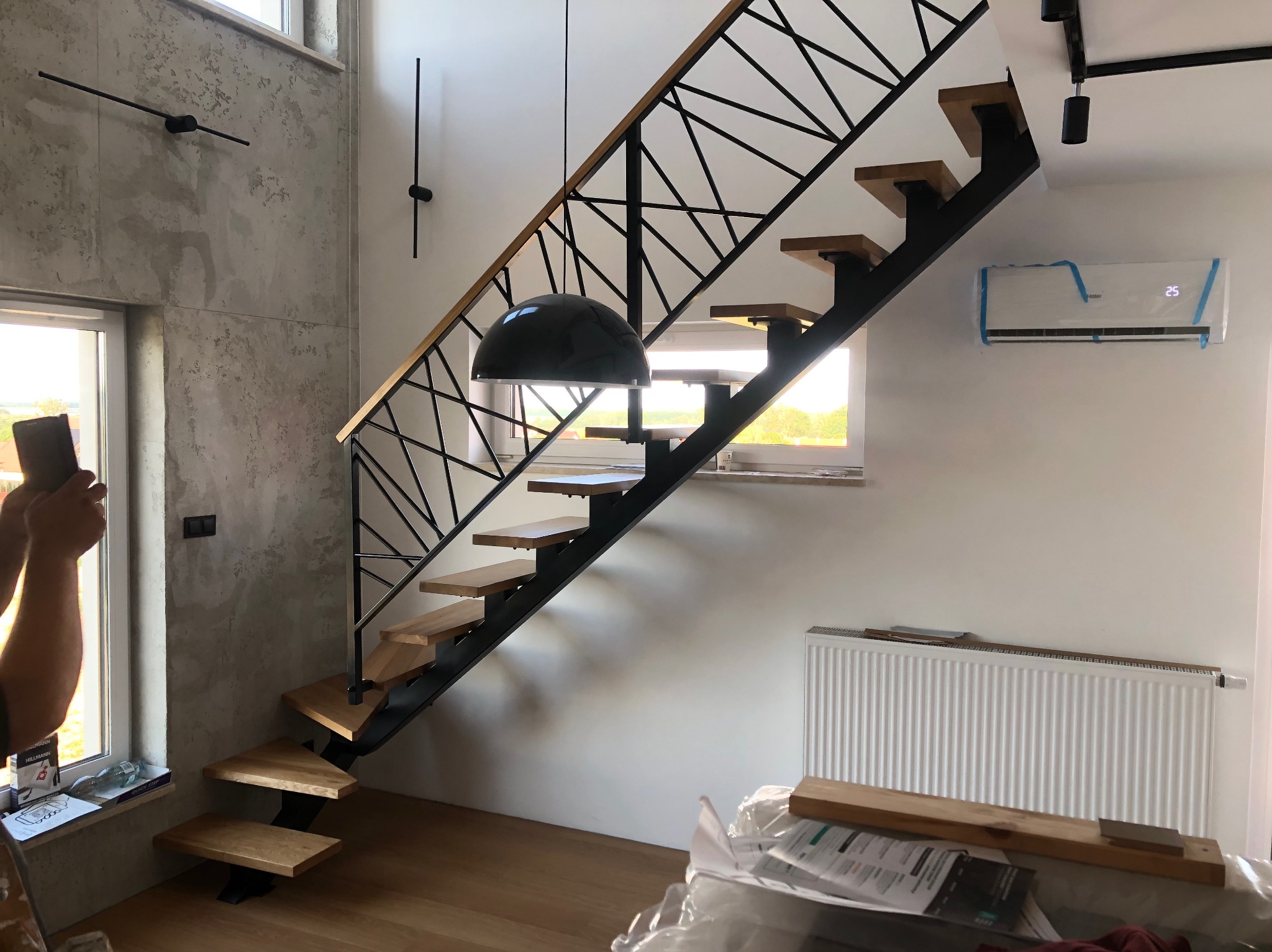 Nowoczesne schody z drewnianymi stopniami i metalową, geometryczną balustradą, oświetlone wiszącą lampą w kształcie kopuły, sfotografowane podczas montażu - widoczne narzędzia i osoba robiąca zdjęcie.