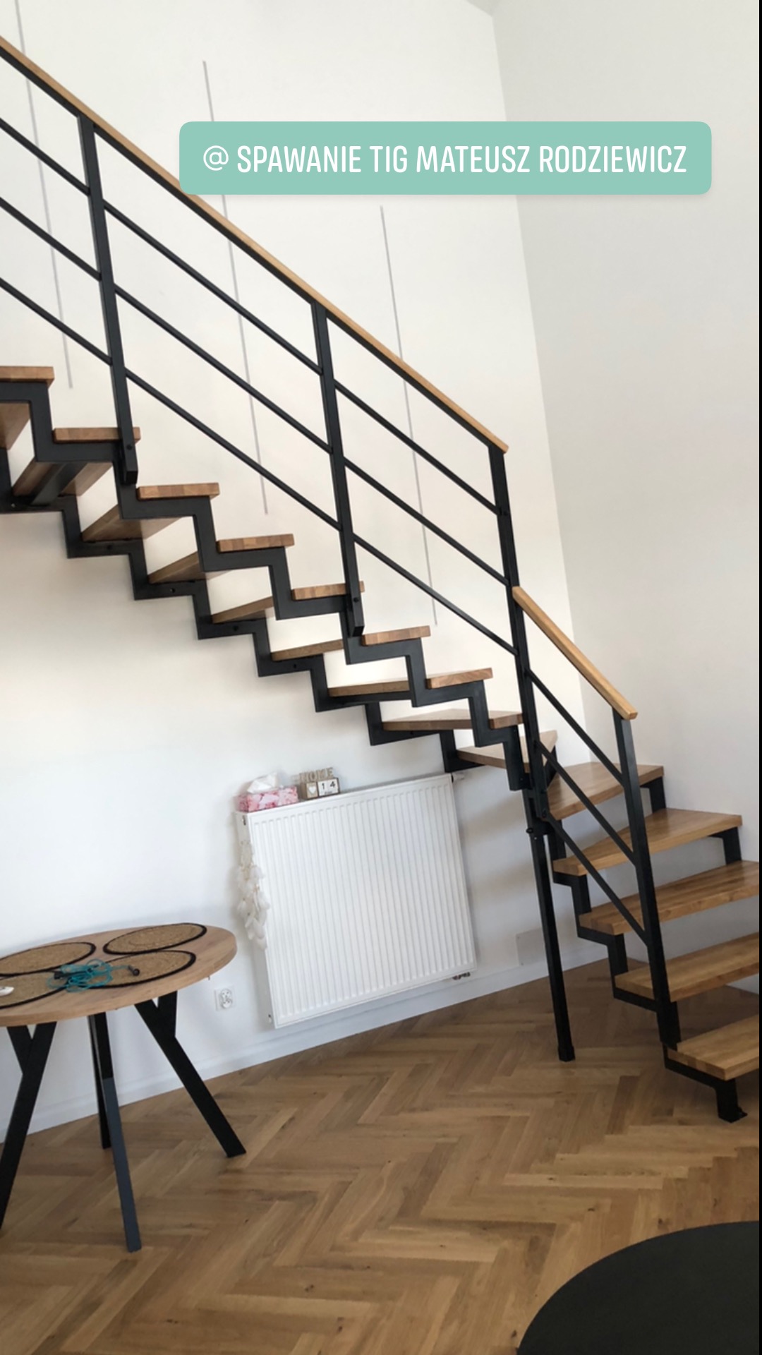 Nowoczesne schody metalowe z drewnianymi stopniami i czarną, minimalistyczną balustradą, widoczne w jasnym wnętrzu z parkietem w jodełkę.