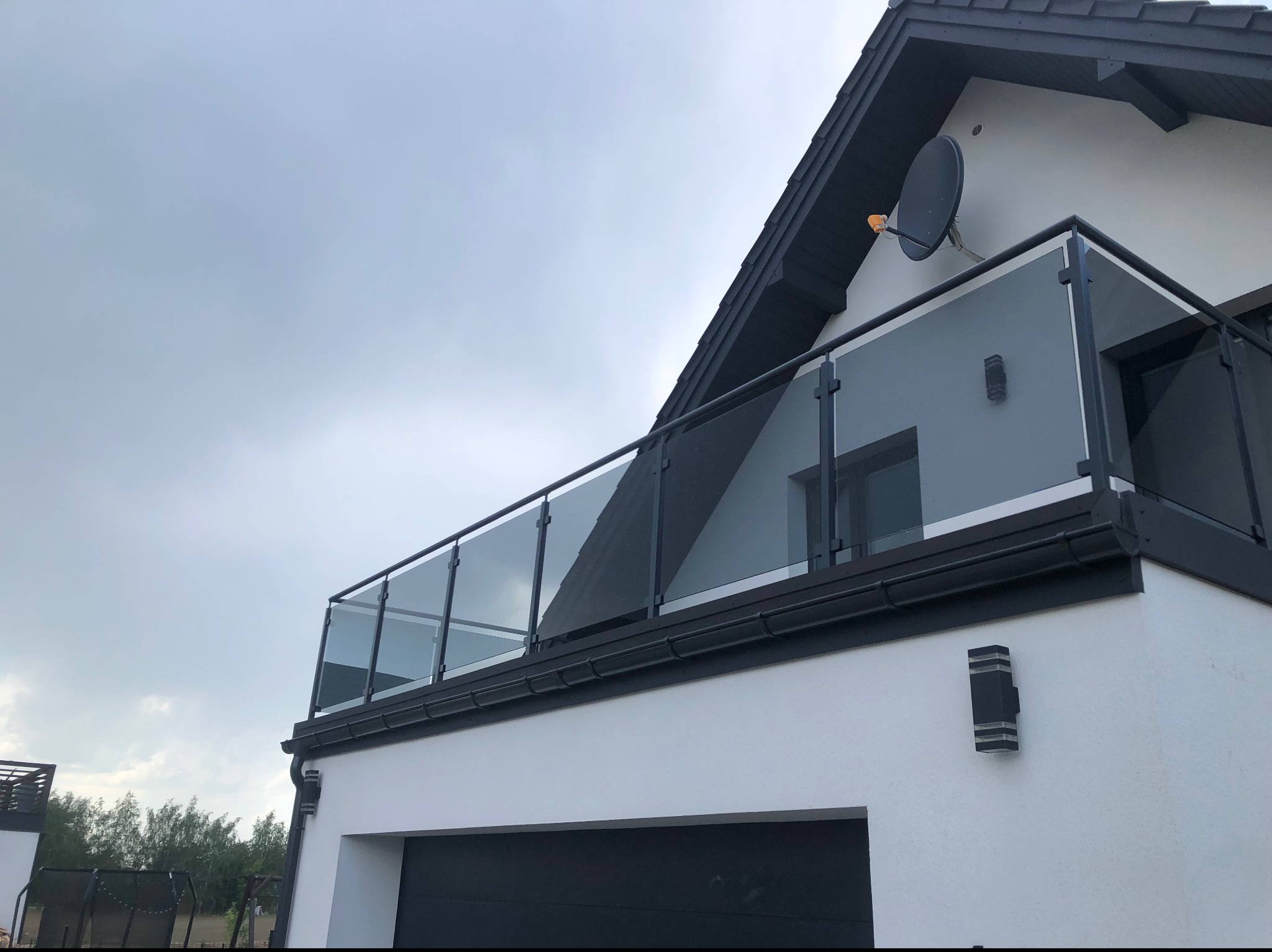 Nowoczesna balustrada szklana z czarnymi elementami mocującymi na balkonie domu jednorodzinnego, widok z dołu w pochmurny dzień.