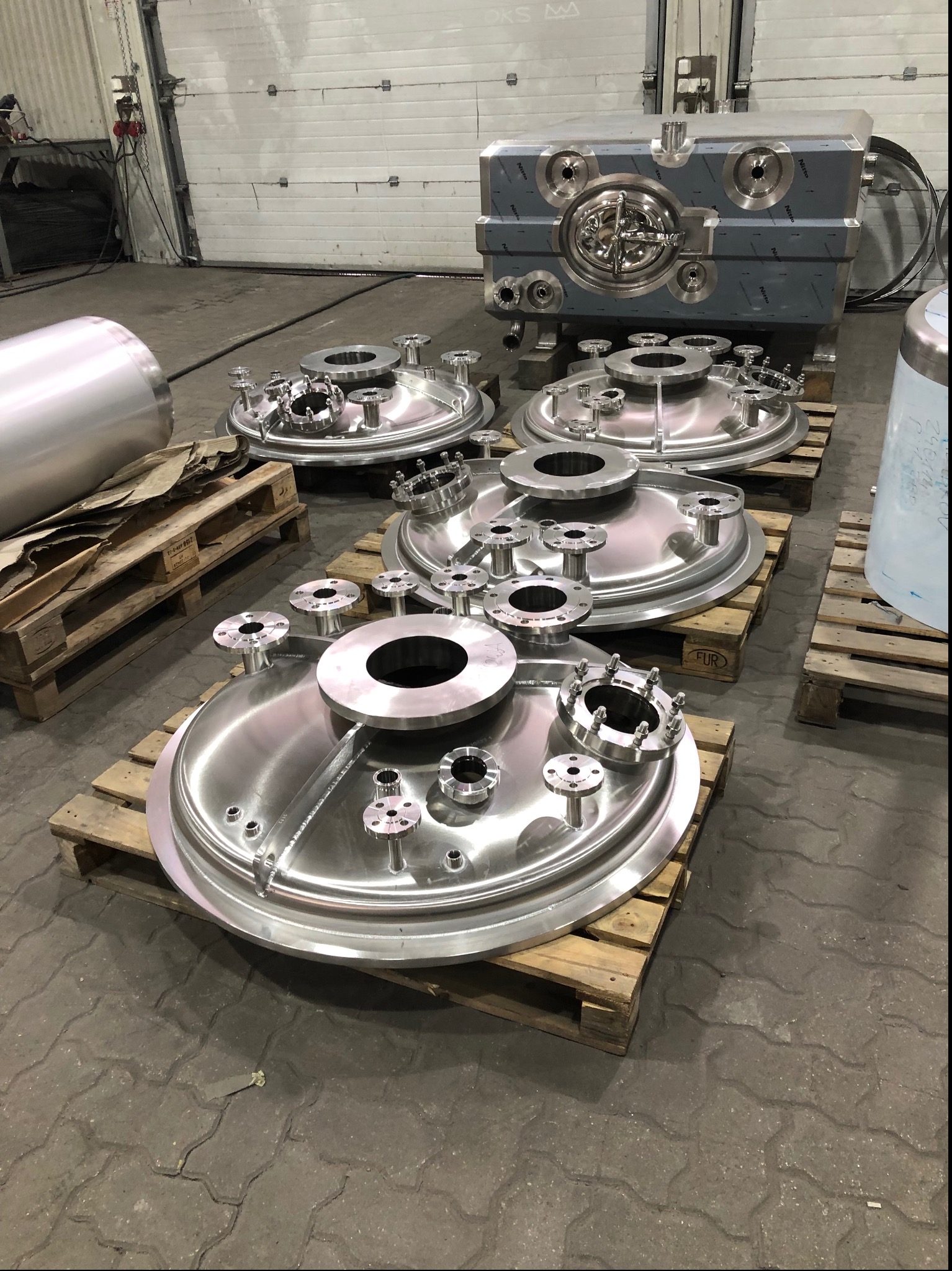 Polerowane, stalowe elementy zbiorników ciśnieniowych na drewnianych paletach w hali produkcyjnej, widoczne różne rozmiary i konfiguracje przyłączy.