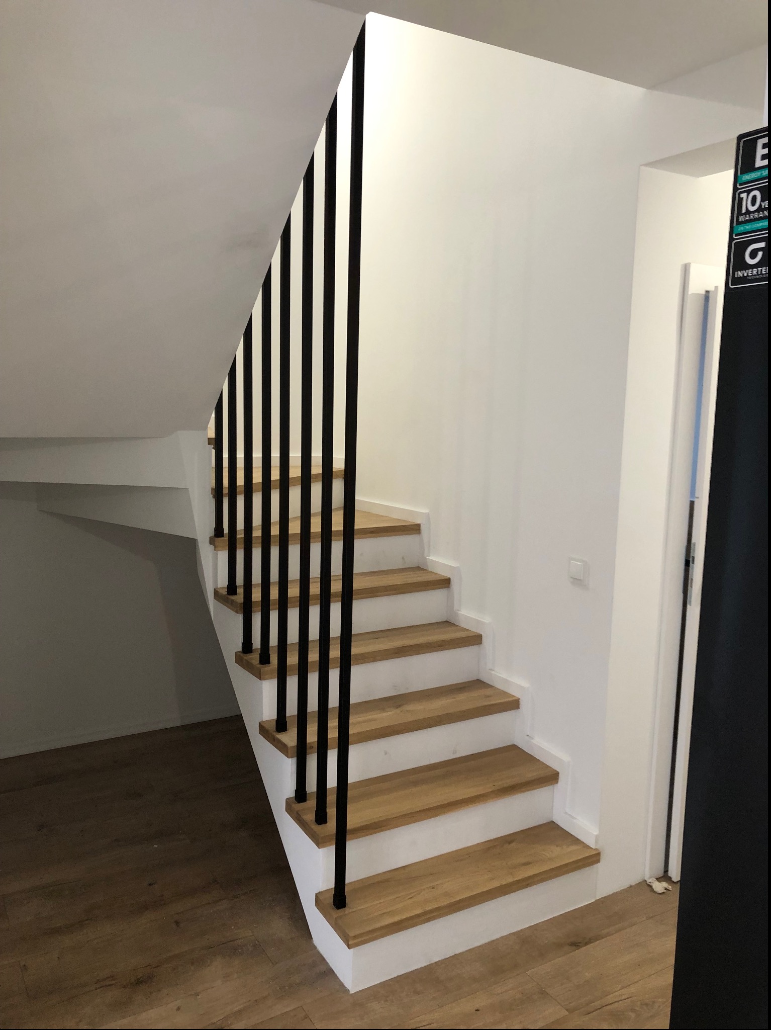 Nowoczesne schody z drewnianymi stopniami i czarnymi, pionowymi elementami balustrady w jasnym wnętrzu.