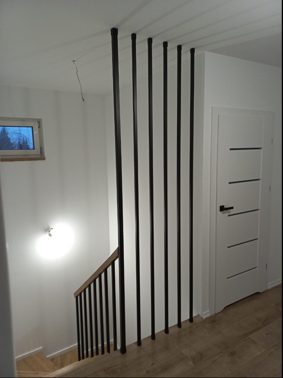 Wnętrze klatki schodowej z drewnianymi schodami i balustradą z czarnych, pionowych prętów, widoczna biała ściana z oknem i białe drzwi z poziomymi wgłębieniami, oświetlenie sufitowe.